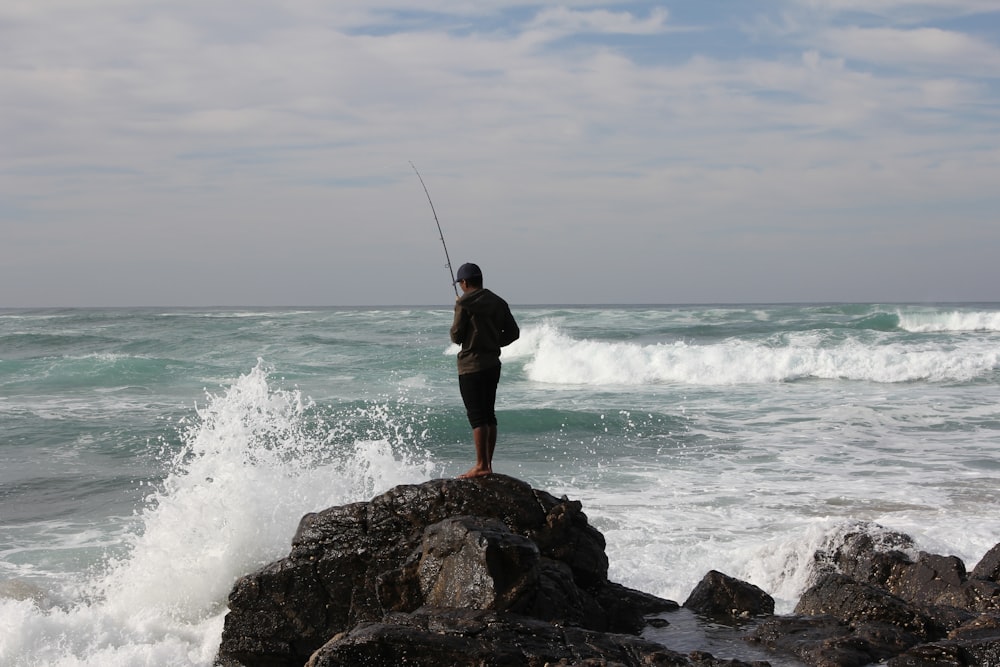 uomo in pantaloncini neri in piedi sulla roccia vicino al mare durante il giorno