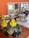 A collection of children's toys and objects are arranged on a textured surface. Bright yellow rubber ducks are placed beside smooth stones and colorful glass gems. In the background, wooden shelves hold various wooden blocks, cylinders, and domino pieces. A decorative, colorful bird is also visible among the items, adding a playful and artistic touch.