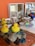 A collection of children's toys and objects are arranged on a textured surface. Bright yellow rubber ducks are placed beside smooth stones and colorful glass gems. In the background, wooden shelves hold various wooden blocks, cylinders, and domino pieces. A decorative, colorful bird is also visible among the items, adding a playful and artistic touch.