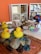 A collection of children's toys and objects are arranged on a textured surface. Bright yellow rubber ducks are placed beside smooth stones and colorful glass gems. In the background, wooden shelves hold various wooden blocks, cylinders, and domino pieces. A decorative, colorful bird is also visible among the items, adding a playful and artistic touch.
