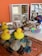 A collection of children's toys and objects are arranged on a textured surface. Bright yellow rubber ducks are placed beside smooth stones and colorful glass gems. In the background, wooden shelves hold various wooden blocks, cylinders, and domino pieces. A decorative, colorful bird is also visible among the items, adding a playful and artistic touch.