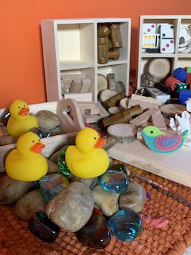 A collection of children's toys and objects are arranged on a textured surface. Bright yellow rubber ducks are placed beside smooth stones and colorful glass gems. In the background, wooden shelves hold various wooden blocks, cylinders, and domino pieces. A decorative, colorful bird is also visible among the items, adding a playful and artistic touch.