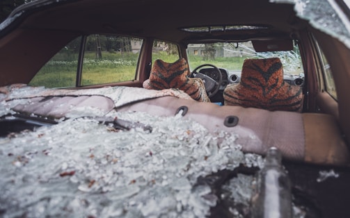 The interior of a car is shown with shattered glass scattered across the backseat and rear deck. Two car seats are covered with an orange and black patterned fabric resembling tiger stripes. The windows have been smashed, and through the windshield view, a grassy area with some trees is visible.