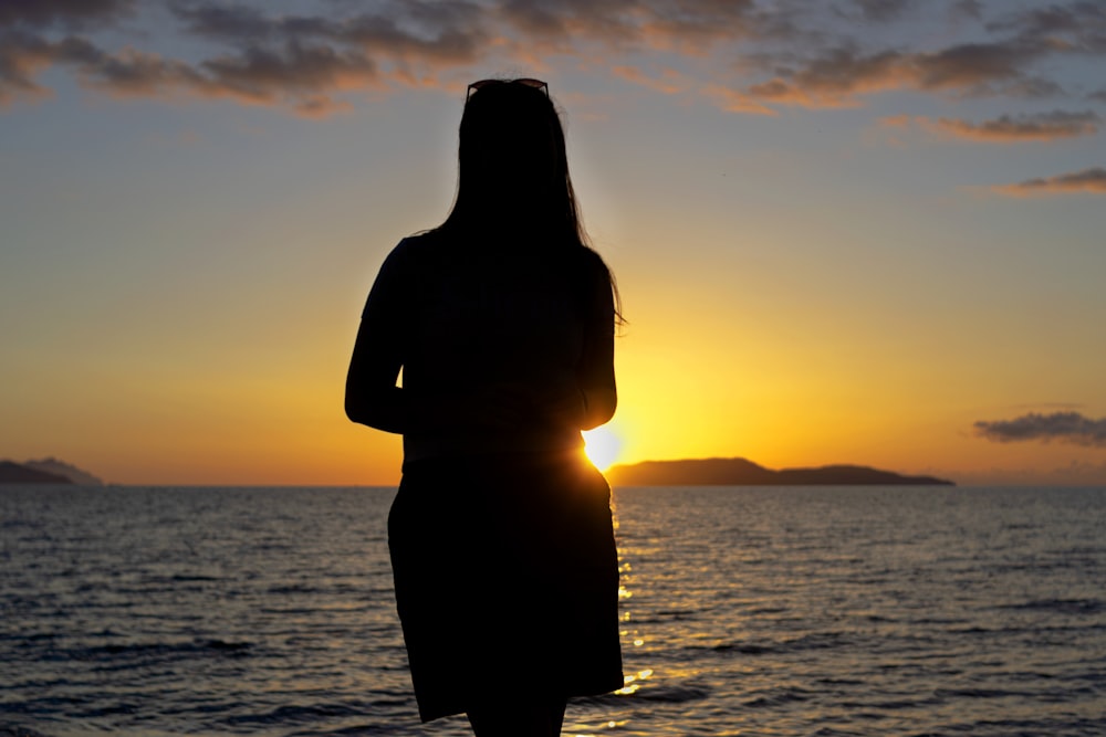 silhouette of woman standing on sea shore during sunset