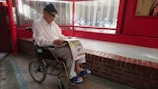An elderly person wearing a white shirt and patterned pants sits in a wheelchair, reading a newspaper. The setting appears to be indoors with red walls and a large window. The floor is made of brick and tiles, with a red brick ledge behind the person.