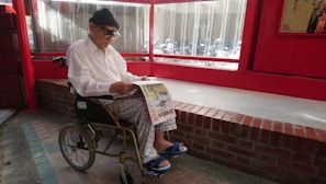 An elderly person wearing a white shirt and patterned pants sits in a wheelchair, reading a newspaper. The setting appears to be indoors with red walls and a large window. The floor is made of brick and tiles, with a red brick ledge behind the person.