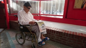 An elderly person wearing a white shirt and patterned pants sits in a wheelchair, reading a newspaper. The setting appears to be indoors with red walls and a large window. The floor is made of brick and tiles, with a red brick ledge behind the person.