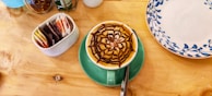 A cup of coffee with an intricate chocolate design on the froth is placed on a green saucer. The coffee is on a wooden table alongside a ceramic container holding various tea bags and a decorative plate with blue floral patterns.