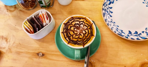 A cup of coffee with an intricate chocolate design on the froth is placed on a green saucer. The coffee is on a wooden table alongside a ceramic container holding various tea bags and a decorative plate with blue floral patterns.