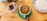 A cup of coffee with an intricate chocolate design on the froth is placed on a green saucer. The coffee is on a wooden table alongside a ceramic container holding various tea bags and a decorative plate with blue floral patterns.