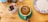 A cup of coffee with an intricate chocolate design on the froth is placed on a green saucer. The coffee is on a wooden table alongside a ceramic container holding various tea bags and a decorative plate with blue floral patterns.