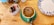 A cup of coffee with an intricate chocolate design on the froth is placed on a green saucer. The coffee is on a wooden table alongside a ceramic container holding various tea bags and a decorative plate with blue floral patterns.