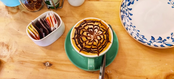 A cup of coffee with an intricate chocolate design on the froth is placed on a green saucer. The coffee is on a wooden table alongside a ceramic container holding various tea bags and a decorative plate with blue floral patterns.