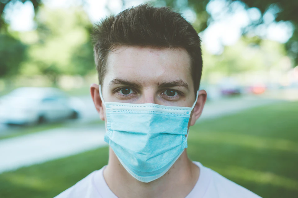 Young man wears blue surgical mask outdoors in front of trees and parked cars