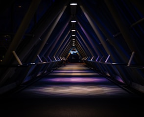A dimly lit, futuristic corridor with a triangular structure, illuminated by overhead lights casting symmetrical shadows and reflections on the floor. The perspective creates a vanishing point at the far end, adding depth to the scene.