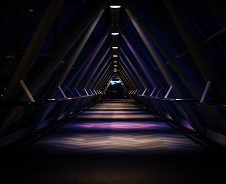 A dimly lit, futuristic corridor with a triangular structure, illuminated by overhead lights casting symmetrical shadows and reflections on the floor. The perspective creates a vanishing point at the far end, adding depth to the scene.