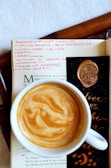 A cup of coffee with a beautifully swirled crema sits atop an open book. The page contains handwritten pink notes, with words like 'paranoid' and 'self-destructive'. A black bookmark decorated with an embossed tree wax seal and gold lettering is partially visible.