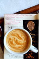 A cup of coffee with a beautifully swirled crema sits atop an open book. The page contains handwritten pink notes, with words like 'paranoid' and 'self-destructive'. A black bookmark decorated with an embossed tree wax seal and gold lettering is partially visible.