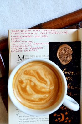 A cup of coffee with a beautifully swirled crema sits atop an open book. The page contains handwritten pink notes, with words like 'paranoid' and 'self-destructive'. A black bookmark decorated with an embossed tree wax seal and gold lettering is partially visible.