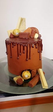 A tall chocolate cake with smooth icing features a dark chocolate drip running down the sides. The top is adorned with a variety of chocolates including Hershey's chocolate bar, Ferrero Rocher, wafers, and other assorted candies. The cake is placed on a glass cake stand.