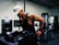 A man wearing a white baseball cap and a black tank top is engaged in a workout at a gym. He has tattoos on his arm and is using gym equipment designed for dips. The background includes more gym equipment and a window, suggesting an indoor gym setting.