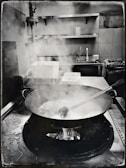 A commercial kitchen setting with a large pan on a gas stove, emitting steam. A metal ladle rests inside the pan. Surrounding the stove are stainless steel surfaces, a sink, and industrial shelves. Water stains are visible on the counter.