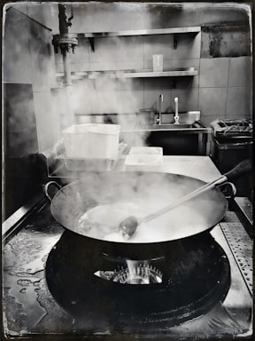 A commercial kitchen setting with a large pan on a gas stove, emitting steam. A metal ladle rests inside the pan. Surrounding the stove are stainless steel surfaces, a sink, and industrial shelves. Water stains are visible on the counter.