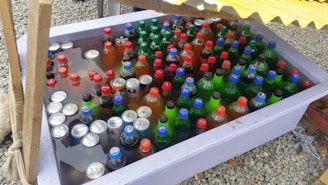 A large plastic container filled with various bottled and canned drinks submerged in water. The bottles and cans have colorful caps and labels, predominantly in red, blue, and green. The container is set on a gravel surface, partially shaded by a yellow corrugated cover.