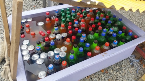 A large plastic container filled with various bottled and canned drinks submerged in water. The bottles and cans have colorful caps and labels, predominantly in red, blue, and green. The container is set on a gravel surface, partially shaded by a yellow corrugated cover.