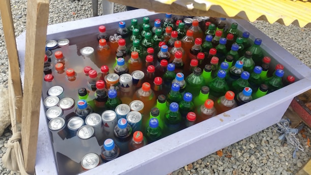 A large plastic container filled with various bottled and canned drinks submerged in water. The bottles and cans have colorful caps and labels, predominantly in red, blue, and green. The container is set on a gravel surface, partially shaded by a yellow corrugated cover.