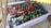A large plastic container filled with various bottled and canned drinks submerged in water. The bottles and cans have colorful caps and labels, predominantly in red, blue, and green. The container is set on a gravel surface, partially shaded by a yellow corrugated cover.