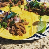Corn tortillas are topped with shredded meat, chopped red onion, and fresh cilantro, served on a plate with slices of lime. The setting is a granite table surface.