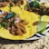 Corn tortillas are topped with shredded meat, chopped red onion, and fresh cilantro, served on a plate with slices of lime. The setting is a granite table surface.