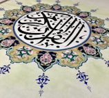 An intricate piece of Islamic art features Arabic calligraphy at its center, surrounded by an ornate floral design. The calligraphy is in bold black lines, creating a striking contrast against a cream background, while the floral elements are rich with details, displaying pink and green hues with blue accents.