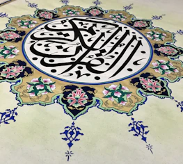 An intricate piece of Islamic art features Arabic calligraphy at its center, surrounded by an ornate floral design. The calligraphy is in bold black lines, creating a striking contrast against a cream background, while the floral elements are rich with details, displaying pink and green hues with blue accents.