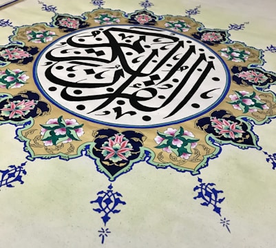 An intricate piece of Islamic art features Arabic calligraphy at its center, surrounded by an ornate floral design. The calligraphy is in bold black lines, creating a striking contrast against a cream background, while the floral elements are rich with details, displaying pink and green hues with blue accents.