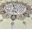 An intricate piece of Islamic art features Arabic calligraphy at its center, surrounded by an ornate floral design. The calligraphy is in bold black lines, creating a striking contrast against a cream background, while the floral elements are rich with details, displaying pink and green hues with blue accents.