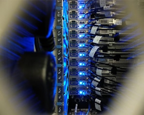 A close-up view of a server rack in a data center, showing rows of servers with blue LED lights glowing. Several network cables are connected to the servers, with labels indicating their connections.