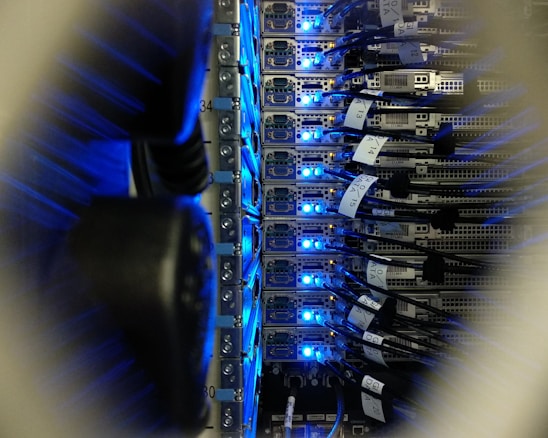 A close-up view of a server rack in a data center, showing rows of servers with blue LED lights glowing. Several network cables are connected to the servers, with labels indicating their connections.