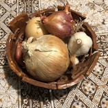 A rustic ceramic bowl holds a variety of onions and a bulb of garlic. The onions include both yellow and red varieties. These vegetables rest on a patterned tablecloth with floral and geometric designs in shades of brown and white.