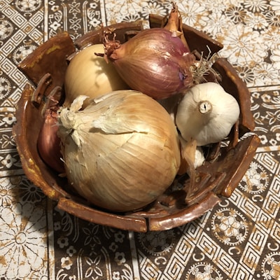 A rustic ceramic bowl holds a variety of onions and a bulb of garlic. The onions include both yellow and red varieties. These vegetables rest on a patterned tablecloth with floral and geometric designs in shades of brown and white.
