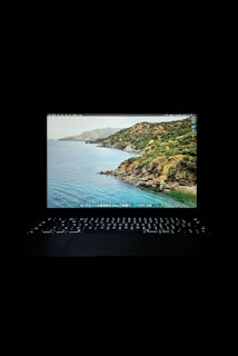 A laptop displaying a scenic wallpaper with a view of a coastline and ocean. The screen is bright against the dark surroundings, and the keyboard is illuminated with a soft glow.