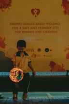 A child is seated in front of a large poster advocating against gender-based violence, with a message promoting safety for women and children. The child holds a lit lantern and is wearing casual clothing. The setting appears to be in a public area with subdued lighting.