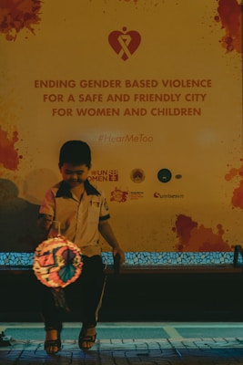 A child is seated in front of a large poster advocating against gender-based violence, with a message promoting safety for women and children. The child holds a lit lantern and is wearing casual clothing. The setting appears to be in a public area with subdued lighting.