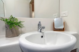 A clean and modern bathroom sink with a chrome faucet is running water. To the side, a small indoor plant in a beige fabric container adds a touch of greenery. A compact rectangular mirror with a wooden frame rests on the counter next to electrical outlets.