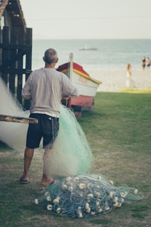 A person stands on a grassy area near the ocean holding a fishing net. In the background, a boat is visible with a sandy beach and the sea stretching out. There are a few people walking on the beach, and another boat floats in the distance on the water.