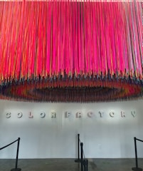 A vibrant display of colorful hanging strings creates a striking overhead installation in front of a white wall. The words 'COLOR FACTORY' are spelled out in 3D letters on the wall beneath the installation, casting distinct shadows. The floor is a polished concrete surface with stanchions guiding a pathway.