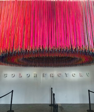A vibrant display of colorful hanging strings creates a striking overhead installation in front of a white wall. The words 'COLOR FACTORY' are spelled out in 3D letters on the wall beneath the installation, casting distinct shadows. The floor is a polished concrete surface with stanchions guiding a pathway.