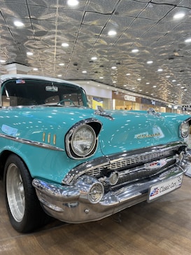 A classic turquoise vintage car is displayed indoors, likely in a showroom or exhibition. The car has a shiny chrome front bumper and a distinct round headlight design from the mid-20th century. There are bright lights on the ceiling, and shopfronts in the background, including a store named Chester.