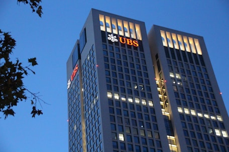 Two tall office buildings with rectangular windows are lit up against a clear evening sky. The UBS logo is prominently displayed on the upper levels of both buildings. Some tree branches are visible on the left side.